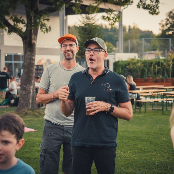 Zwei Männer stehen in einem Park, einer hält ein Getränk, der andere trägt eine Kappe und eine Brille. Ein Kind schaut im Vordergrund zu.
