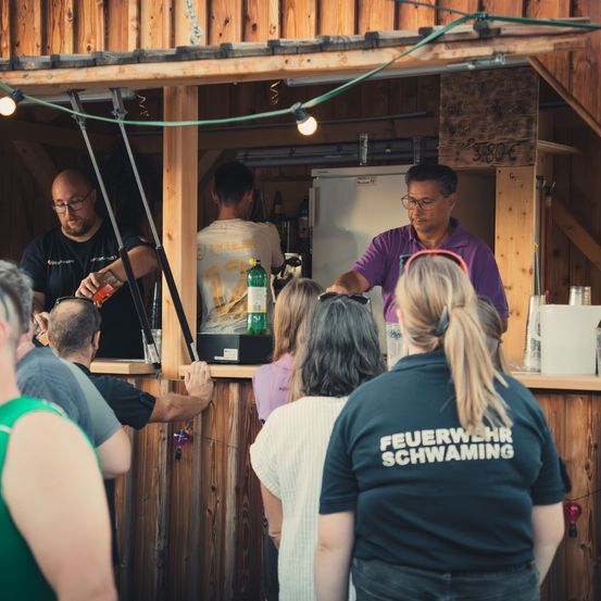 Eine Gruppe von Menschen versammelt sich um eine Holzbar, wobei zwei Männer im Inneren Getränke servieren. Ein Mann schenkt ein Getränk ein, während ein anderer eine Flasche hält. Die Bar ist mit Lichtern dekoriert und hat ein Schild.