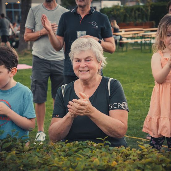 Eine ältere Frau lächelt und klatscht mit einer Gruppe von Menschen in einem Park. Ein Junge und ein Mädchen stehen hinter ihr.