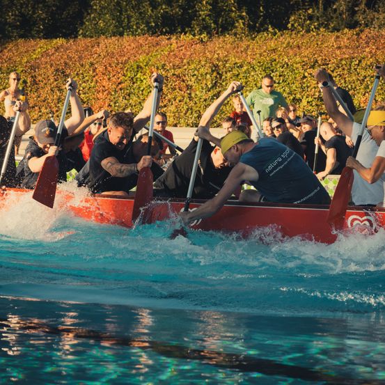 Eine Gruppe von Menschen rudert in einem Pool mit einem roten Boot, wobei Wasserspritzer um sie herum sind.