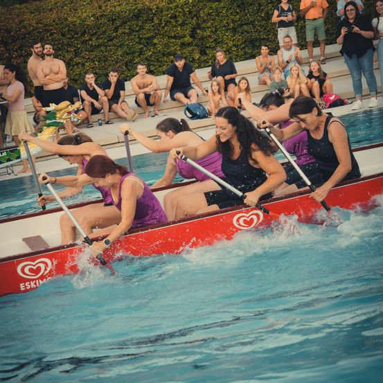 Eine Gruppe von Frauen in lila Tanktops rudert ein rotes Drachenboot im Pool, während Zuschauer von den Rängen zusehen.