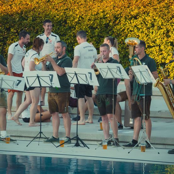 Eine Gruppe von Musikern in grünen Shirts und Shorts spielt Instrumente am Pool, mit anderen, die dahinter stehen.