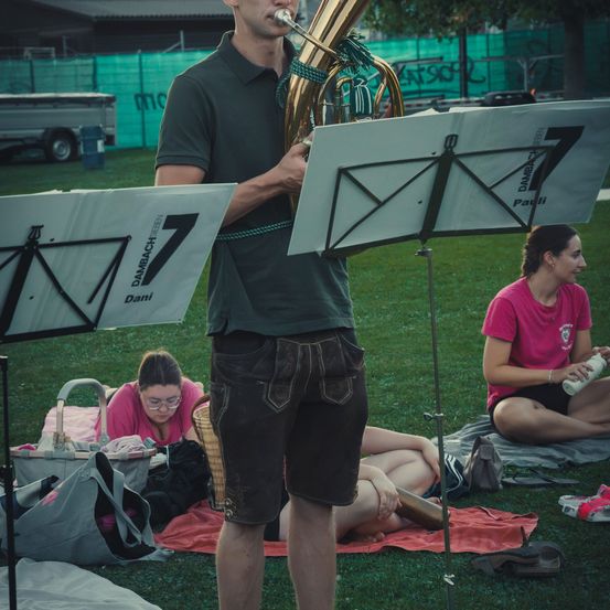 Ein Mann spielt Tuba in einem Park, mit zwei Frauen, die auf dem Gras hinter ihm sitzen. Eine Frau liest und die andere hält eine Flasche.