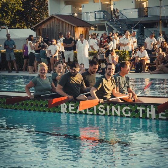 Eine Gruppe von Menschen in grünen Shirts rudert ein Boot im Pool, während Zuschauer von den Seiten zusehen.
