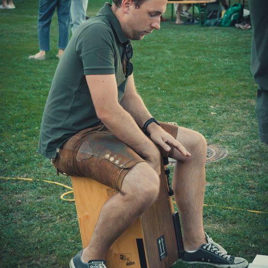 Ein Mann in einem grünen Shirt und braunen Shorts sitzt auf einer Holzkiste in einem Park. Er hat eine Sonnenbrille auf seinem Shirt und schaut nach unten.