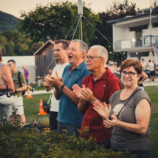 Eine Gruppe von Menschen klatscht und lächelt in einem Park. Ein Gebäude und Bäume sind im Hintergrund.