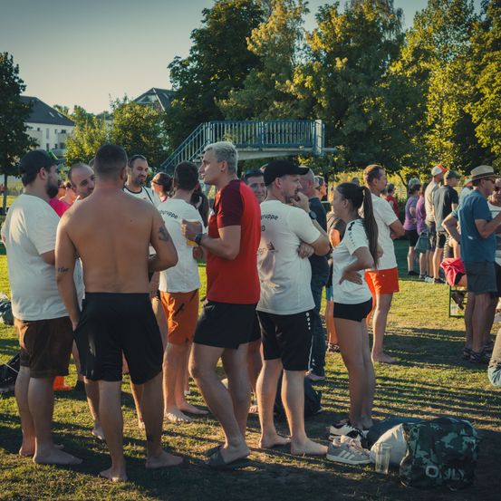 Eine Gruppe von Menschen in einem Park, einige in Sportbekleidung, versammelt sich für eine Veranstaltung. Bäume und Gebäude sind im Hintergrund zu sehen.