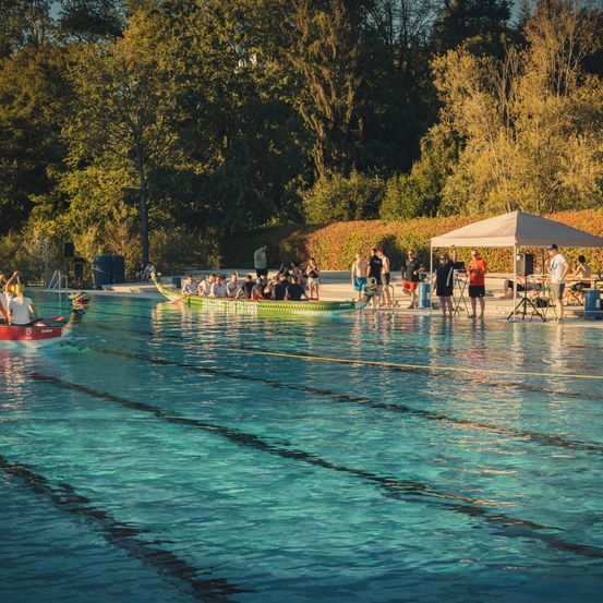 Zwei Drachenboote treten in einem Pool gegeneinander an, während die Leute anfeuern. Bäume und ein Zelt sind im Hintergrund zu sehen.