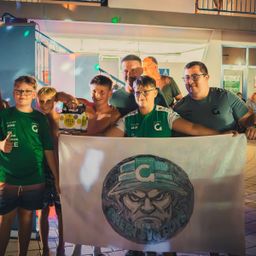 Eine Gruppe von Menschen in grünen Shirts hält ein Banner mit einer Zeichnung eines Soldaten und den Worten 'Green Beret Boys'. Sie posieren für ein Foto vor einem Gebäude.