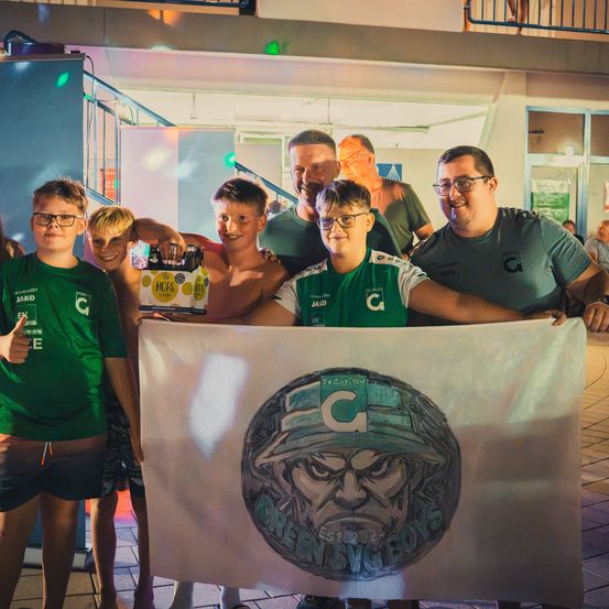 Eine Gruppe von Menschen in grünen Shirts hält ein Banner mit einer Zeichnung eines Soldaten und den Worten 'Green Beret Boys'. Sie posieren für ein Foto vor einem Gebäude.