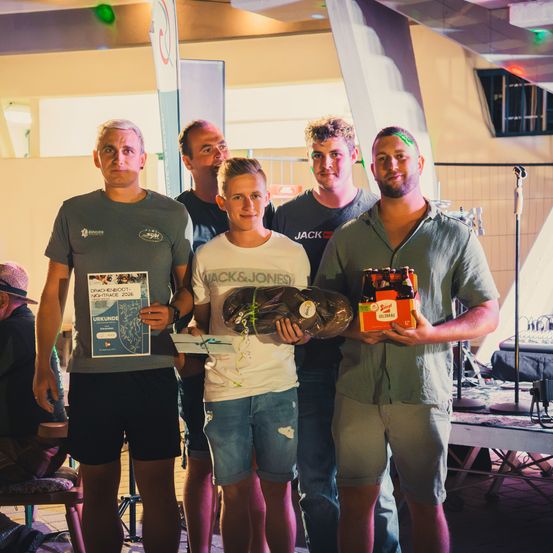 Fünf Männer stehen zusammen, einer hält einen Pokal und ein anderer eine Kiste Bier. Sie lächeln und scheinen eine Veranstaltung zu feiern.