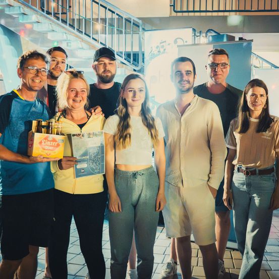 Eine Gruppe von Menschen steht zusammen, lächelt und eine Person hält eine Kiste Bier. Sie befinden sich in einem Raum mit einer Treppe und einer blauen Wand.