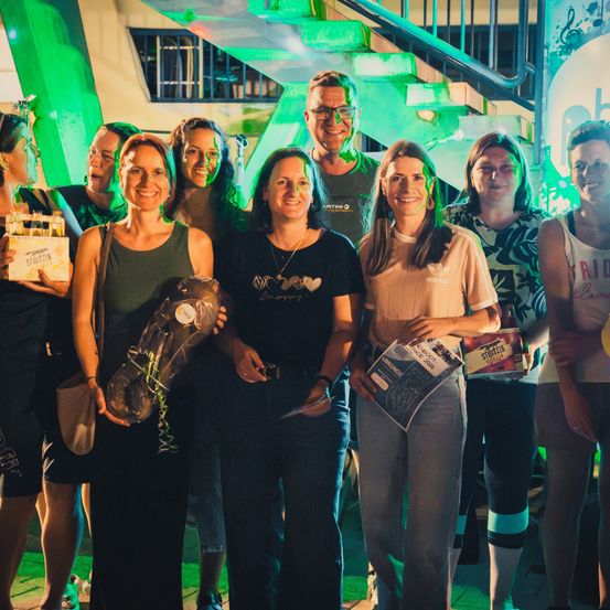 Eine Gruppe von Menschen posiert für ein Foto in einem grün beleuchteten Raum, einer hält einen Pokal und andere halten Getränke.