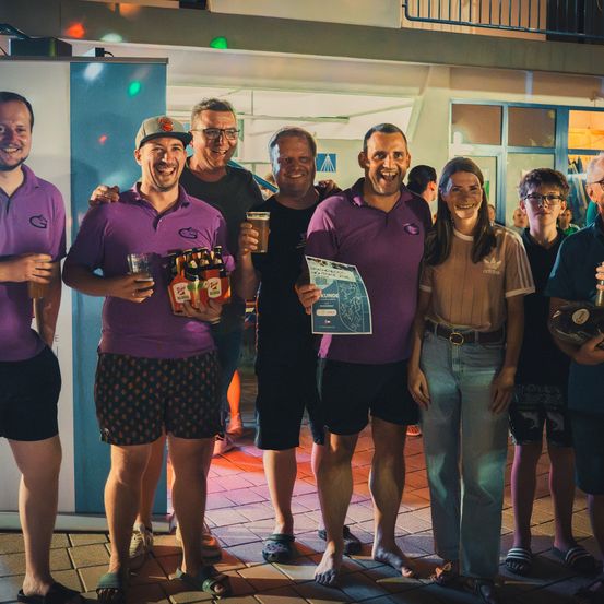 Eine Gruppe von Menschen in lila Hemden, die lächeln und Getränke in der Hand halten, stehen in einer festlichen Umgebung zusammen. Ein Mann hält eine Karte und eine Bierkiste.