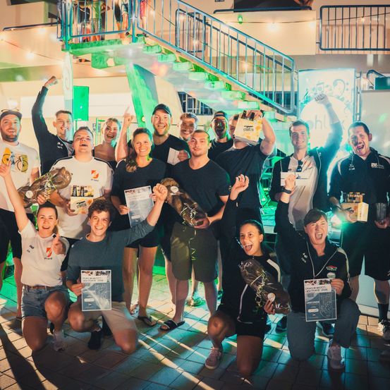 Eine Gruppe von Menschen posiert für ein Foto, hält Trophäen und Getränke in der Hand, mit einer Treppe und grünen Lichtern im Hintergrund.