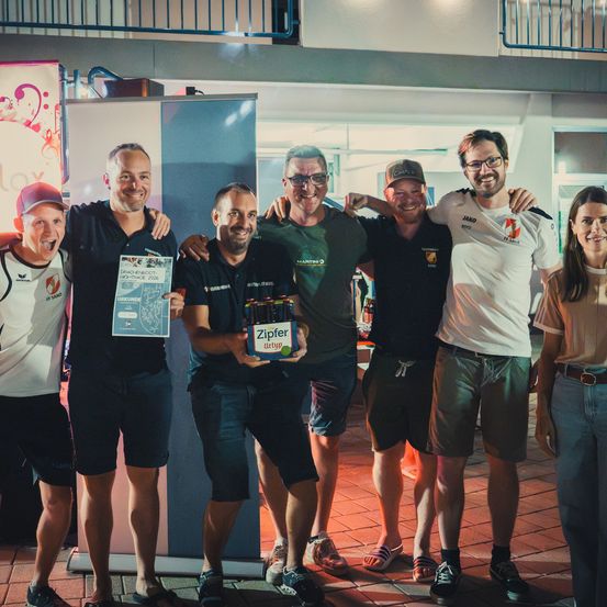 Eine Gruppe von Männern und Frauen posiert für ein Foto, lächelt und hält eine Kiste Bier. Sie stehen auf einem Ziegelboden und ein Mann hält eine Urkunde.