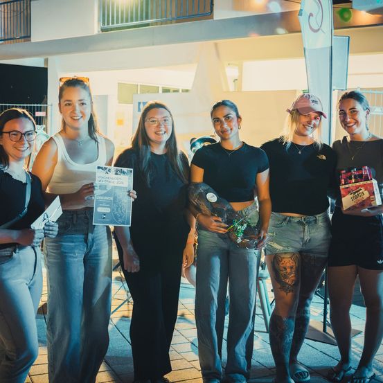 Fünf Frauen stehen bei Nacht zusammen und lächeln für ein Foto. Sie halten verschiedene Gegenstände, darunter ein Zertifikat und eine Getränkekiste. Sie sind lässig gekleidet, eine Frau trägt eine Mütze und eine andere hat ein sichtbares Tattoo.