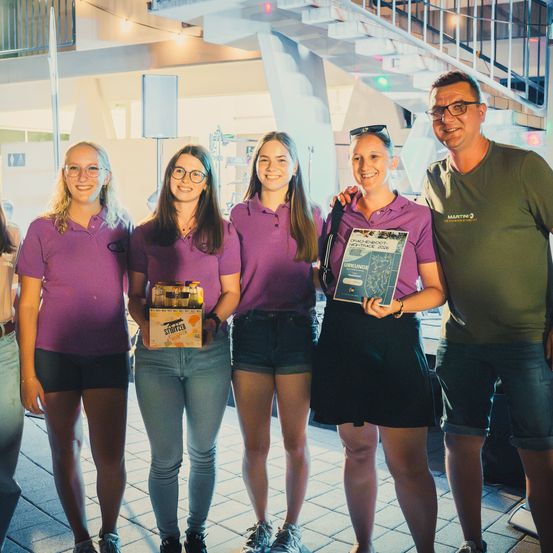 Fünf Personen in lila Poloshirts stehen lächelnd zusammen. Eine Frau hält eine Bierkiste und eine andere ein Zertifikat. Sie befinden sich in einem modernen Gebäude mit einer Treppe im Hintergrund.