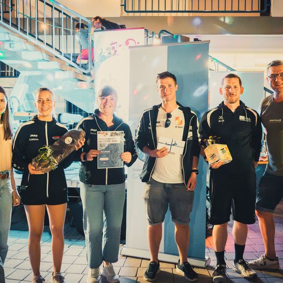 Fünf Personen stehen zusammen und halten Trophäen und Geschenke vor einem blauen Banner. Zwei Frauen halten Blumen und ein Zertifikat, während die Männer Boxen halten. Sie lächeln und scheinen zu feiern.