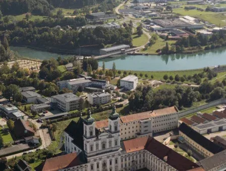 Luftaufnahme einer Stadt mit einem Fluss, Gebäuden und Bäumen. Die Stadt hat eine Kirche, Tennisplätze und ein großes Gebäude. Der Fluss fließt durch die Stadt.