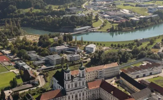 Luftaufnahme einer Stadt mit einem Fluss, Gebäuden und Bäumen. Die Stadt hat eine Kirche, Tennisplätze und ein großes Gebäude. Der Fluss fließt durch die Stadt.