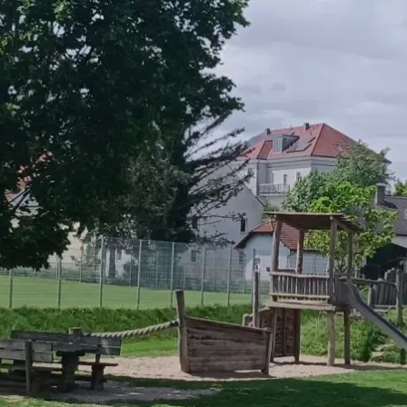 Ein Kinderspielplatz mit Rutsche, Klettergerüst und Bänken unter einem Baum. Ein Zaun umschließt den Spielplatz mit Häusern und einem Feld im Hintergrund.