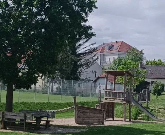 Ein Kinderspielplatz mit Rutsche, Klettergerüst und Bänken unter einem Baum. Ein Zaun umschließt den Spielplatz mit Häusern und einem Feld im Hintergrund.