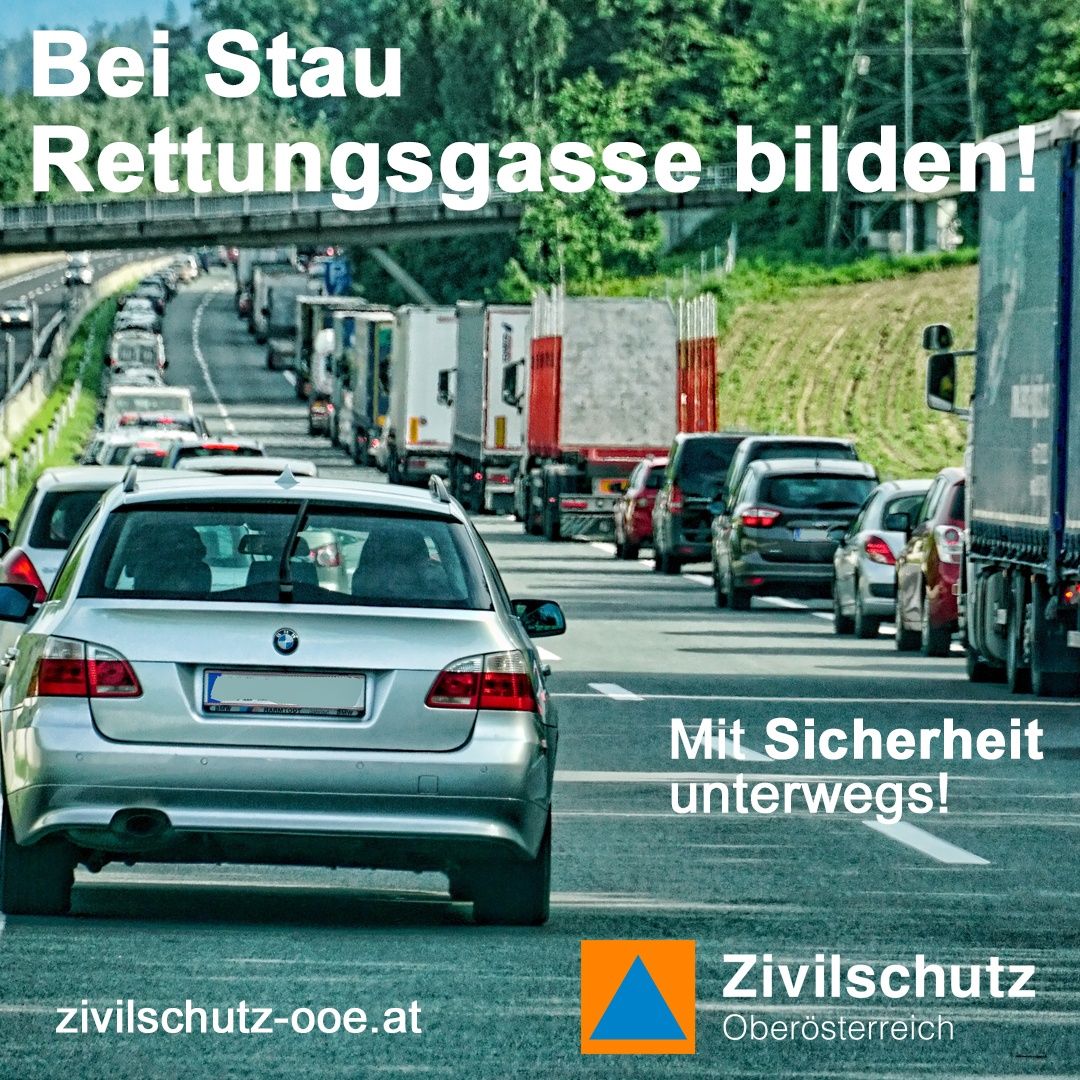Eine Autobahn mit mehreren Spuren, gefüllt mit Autos und Lastwagen. Die Straße hat eine Brücke darüber mit dem Text 'Bei Stau Rettungsgasse bilden!' und einem Logo für Zivilschutz Oberosterreich. Die Autos bewegen sich langsam, und die Straße ist von Bäumen und Gras umgeben.