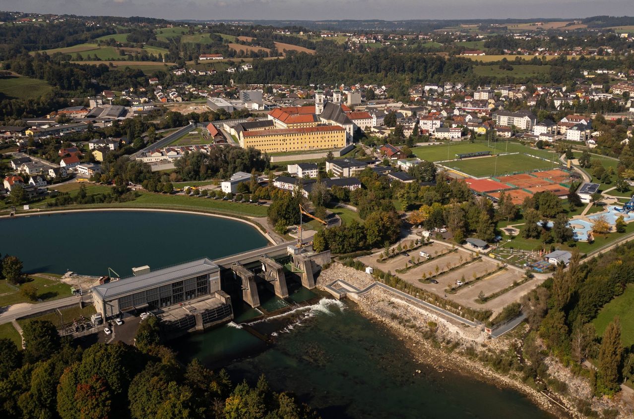 Ein Luftbild einer Stadt mit einem Fluss, einem Damm und einem großen Gebäude. Die Stadt ist von Bäumen umgeben und hat ein großes Feld.