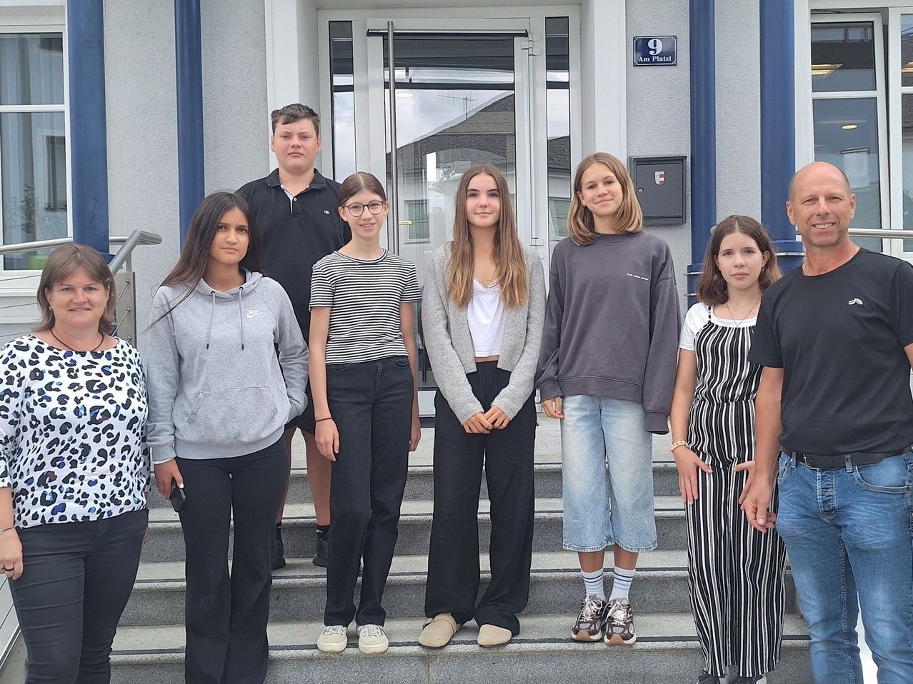 Eine Gruppe junger Menschen, wahrscheinlich Schüler, steht auf den Stufen eines Gebäudes. Sie sind lässig gekleidet und scheinen für ein Foto zu posieren. Das Gebäude hat eine Glastür und ein Schild mit der Aufschrift 'Am Platz 9'.