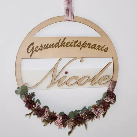 Ein dekorativer Kranz mit dem Namen Nicole hängt an einer weißen Wand. Der Kranz hat ein Band und einen Kranz aus getrockneten Blumen. Der Kranz ist aus Holz.