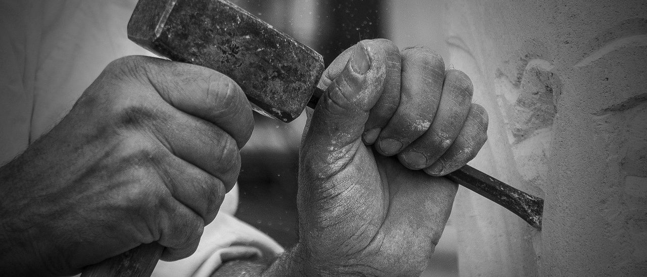 Ein Nahaufnahme von Händen, die einen Hammer halten und an einem Stein arbeiten. Die Hände sind mit Staub bedeckt.