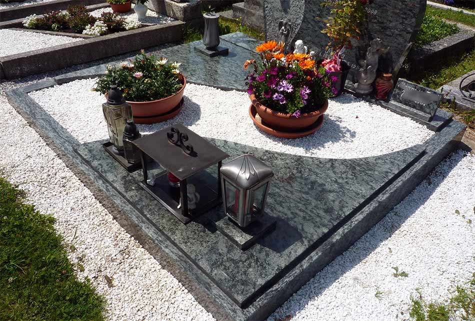 Ein grüner Grabstein ist mit weißem Kies verziert. Er hat einen kleinen Altar mit einer Vase und zwei Lampen. Zwei Blumentöpfe befinden sich auf beiden Seiten.