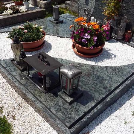 Ein grüner Grabstein ist mit weißem Kies verziert. Er hat einen kleinen Altar mit einer Vase und zwei Lampen. Zwei Blumentöpfe befinden sich auf beiden Seiten.