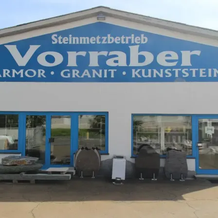 Das Vorraber Steinmetzbetrieb ist spezialisiert auf Panzerung, Granit und Kunststein. Das Geschäft hat blaue Türen, ein großes Schild und Steindisplays draußen.