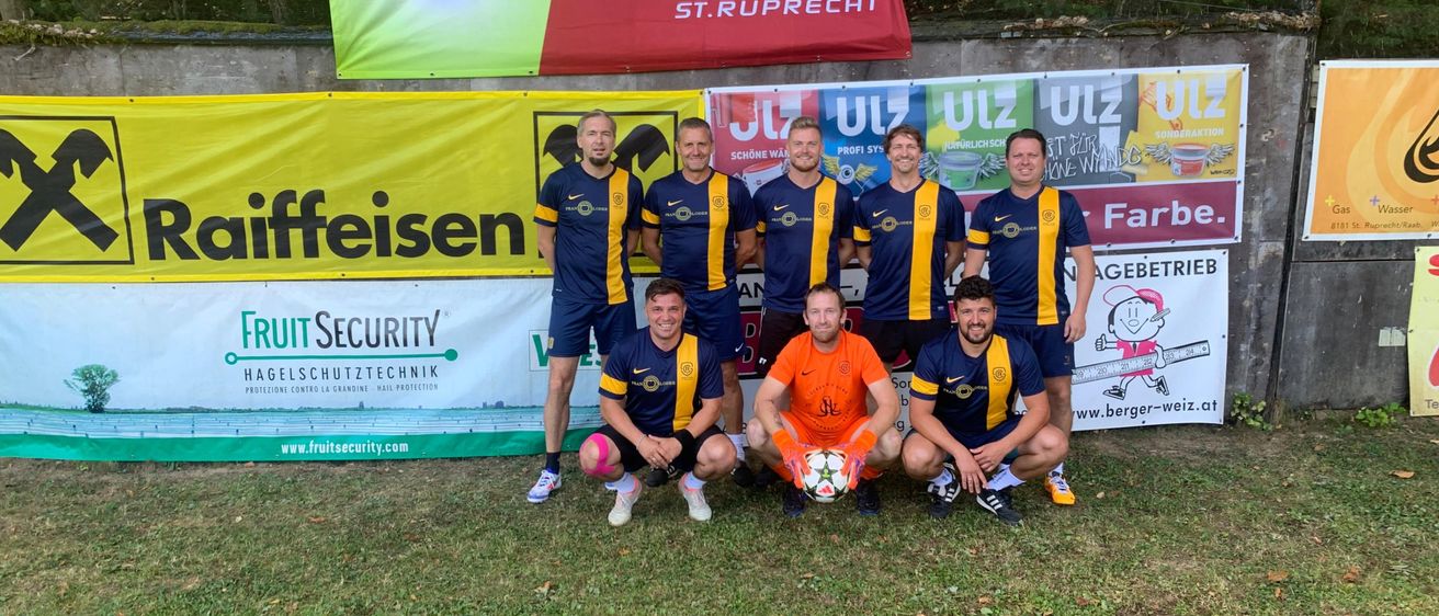 Eine Gruppe von Fußballspielern in blauen und orangefarbenen Trikots posiert für ein Foto mit einem Fußball vor einer Wand mit Bannern.