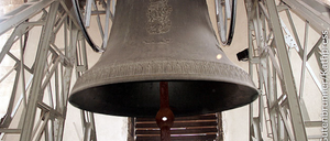 Eine große Glocke hängt von der Decke eines Kirchturms. Die Glocke ist grau und hat ein Design darauf. Die Glocke ist an einer Metallstruktur mit einer Leiter befestigt.
