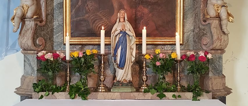 Eine Statue der Jungfrau Maria steht auf einem Marmoraltar, geschmückt mit Blumen und brennenden Kerzen. Der Altar ist von Grünpflanzen umgeben und trägt ein Banner mit einer Botschaft auf Deutsch.