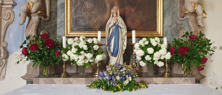 Ein Altar mit einer Statue der Maria, umgeben von weißen und roten Blumen. Hinter dem Altar stehen zwei Cherub-Statuen und ein gerahmtes Gemälde. Der Altar ist mit einem weißen Tuch mit den Worten 'Heilige Maria, bitte für uns' bedeckt.
