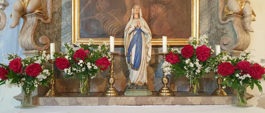 Eine Statue von Maria steht auf einem Altar mit Kerzen und Blumen. Ein Gemälde befindet sich hinter der Statue. Der Altar hat ein weißes Tuch mit rotem Text.