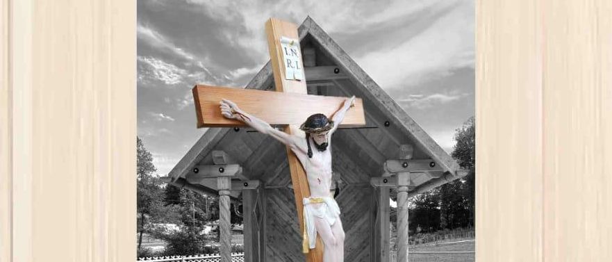 Ein Schwarz-Weiß-Foto eines Kruzifixes vor einer Kapelle mit einem hölzernen Hintergrund. Die Bildunterschrift lautet: 'Nach ca. 170 Jahren Wanderung ist das Steirer-Hauskreuz wieder in seiner Heimat.'