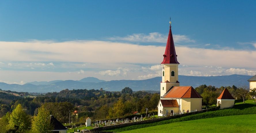 Eine friedliche ländliche Landschaft mit einer kleinen Kirche mit einem roten Turm vor dem Hintergrund von Bergen und grünen Hügeln unter einem teilweise bewölkten Himmel.