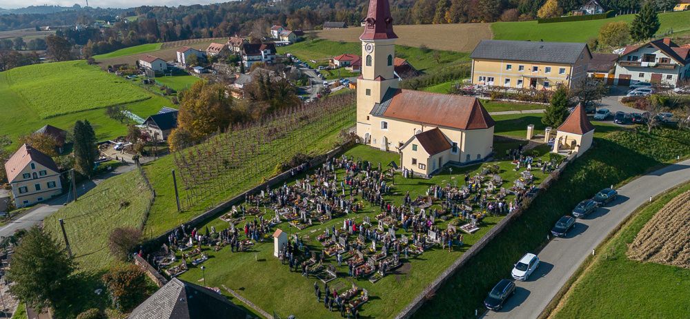 Ein Luftbild einer Kirche mit Friedhof, umgeben von Autos und einem kleinen Dorf unter blauem Himmel.