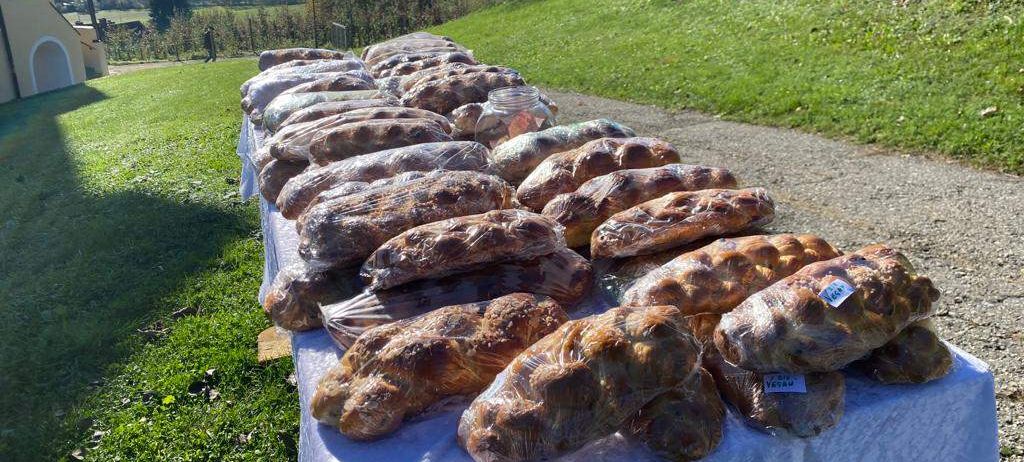 Ein langer Tisch, der mit einem weißen Tuch bedeckt ist, hält zahlreiche Brote, die in Plastik eingewickelt sind. Die Umgebung ist im Freien, mit einem grasbewachsenen Hügel und Bäumen im Hintergrund.