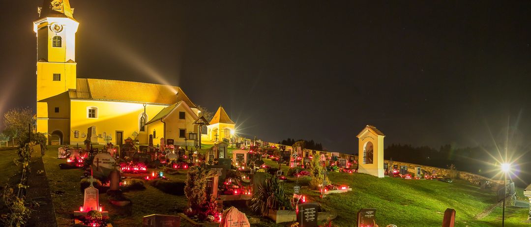 Ein nächtlicher Blick auf einen Friedhof, dessen Grabsteine von roten Kerzen beleuchtet sind, umgeben von einem gepflegten grünen Rasen. Im Hintergrund steht ein gelbes Gebäude mit einem Turm.
