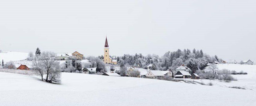 Eine verschneite Dorflandschaft mit einer Kirche und mehreren schneebedeckten Häusern, umgeben von Kiefern und einem klaren Himmel.