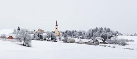 Eine verschneite Dorflandschaft mit einer Kirche und mehreren schneebedeckten Häusern, umgeben von Kiefern und einem klaren Himmel.