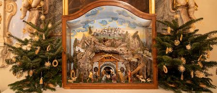 Eine Krippe in einem braunen Holzrahmen wird vor einem großen Gemälde ausgestellt, mit zwei geschmückten Weihnachtsbäumen auf beiden Seiten.
