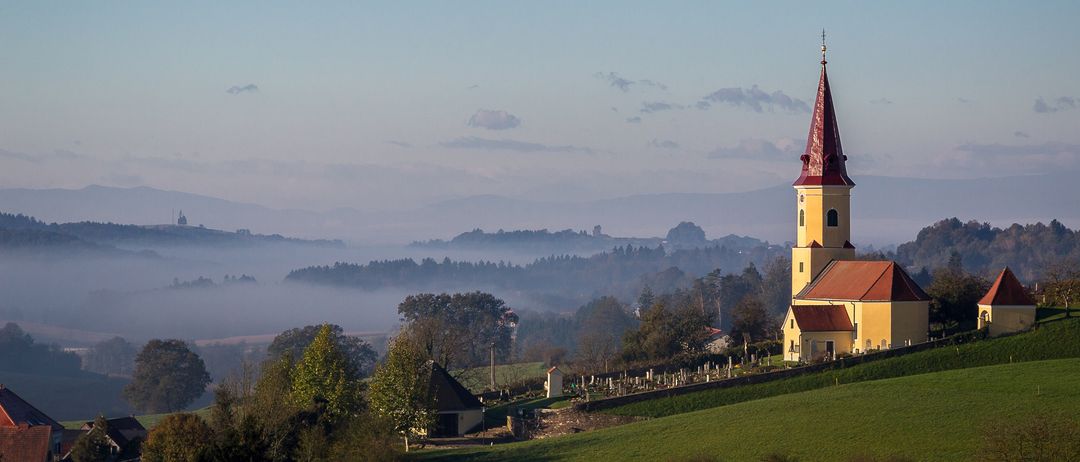 Eine kleine Kirche mit rotem Dach und Turm steht auf einem Hügel, umgeben von Nebel und Bäumen. Der Himmel ist klar und blau.