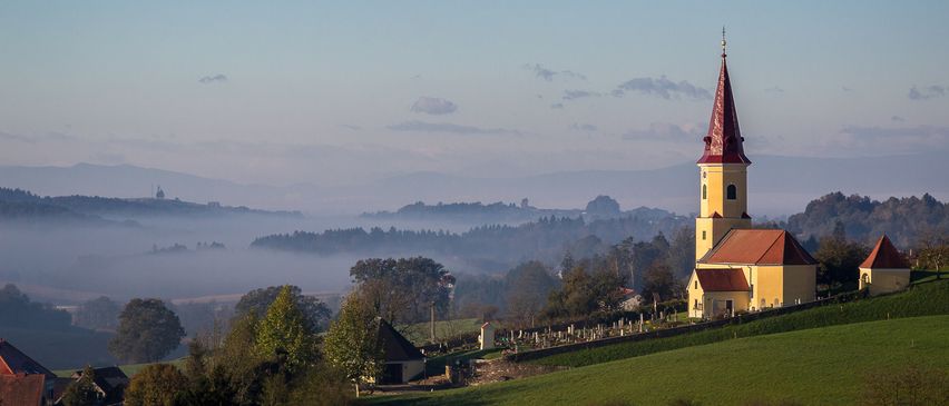 Eine kleine Kirche mit rotem Dach und Turm steht auf einem Hügel, umgeben von Nebel und Bäumen. Der Himmel ist klar und blau.